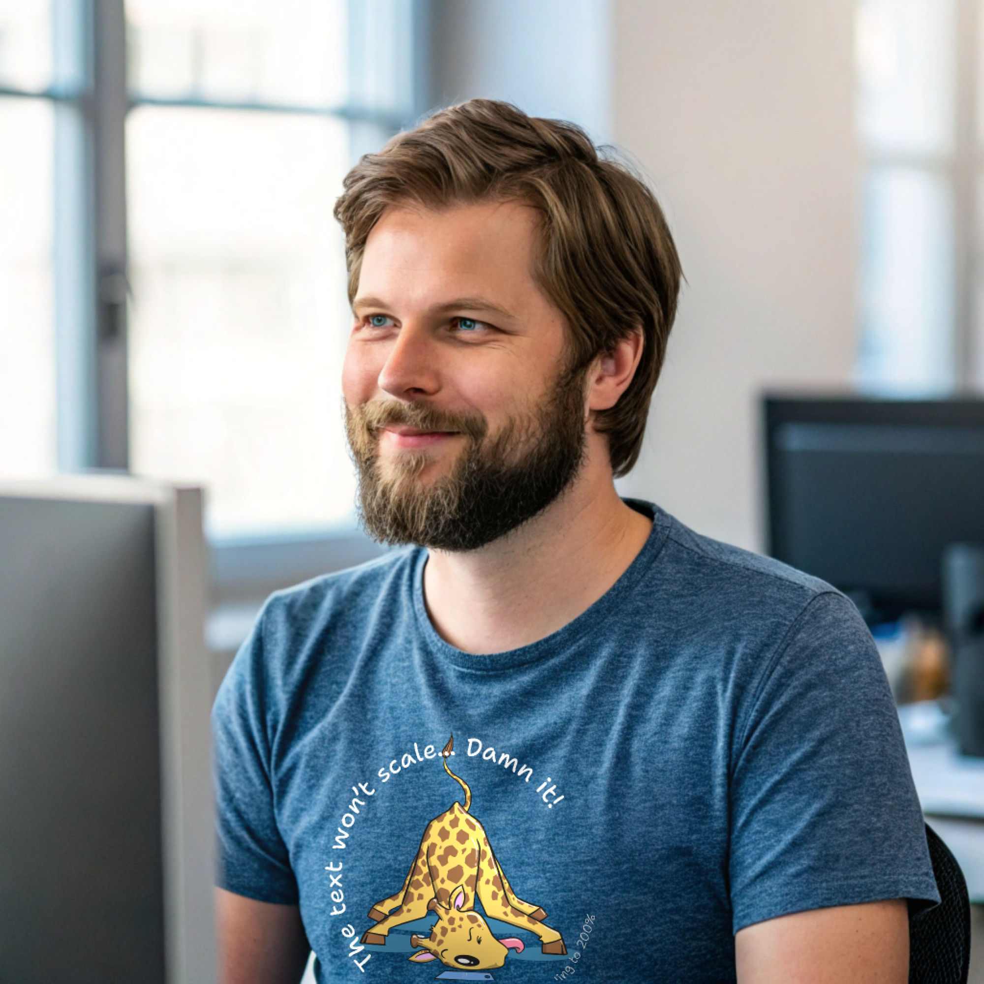 Man at an office desk wearing a blue t-shirt with a cartoon giraffe and the text 'The text won't scale... Damn it!' referencing zooming to 200%