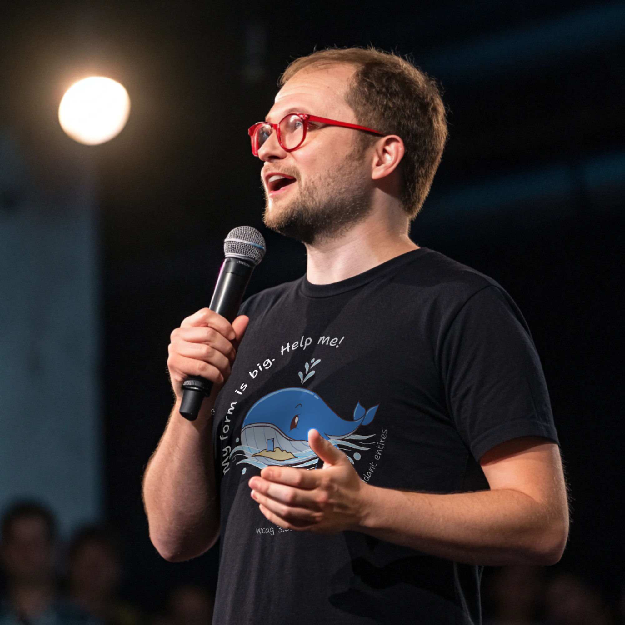 Speaker on stage wearing a black t-shirt with a cartoon whale and the text 'My form is big. Help me!' referencing WCAG 3.3